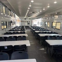 Interior of a long trailer with rows of white tables and black chairs, possibly a temporary workspace.
